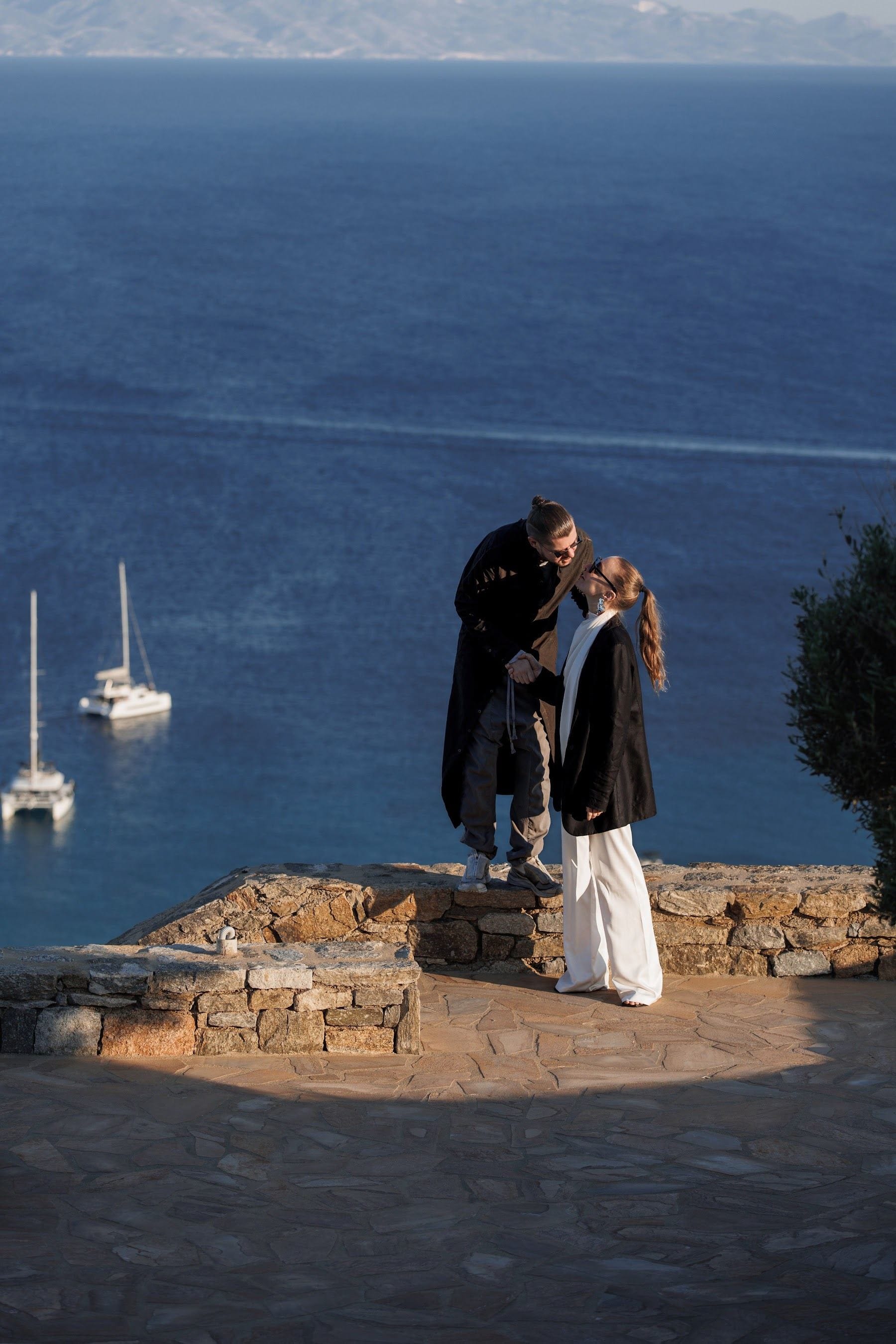 Two guests on a stone terrace above a dark Aegean sea